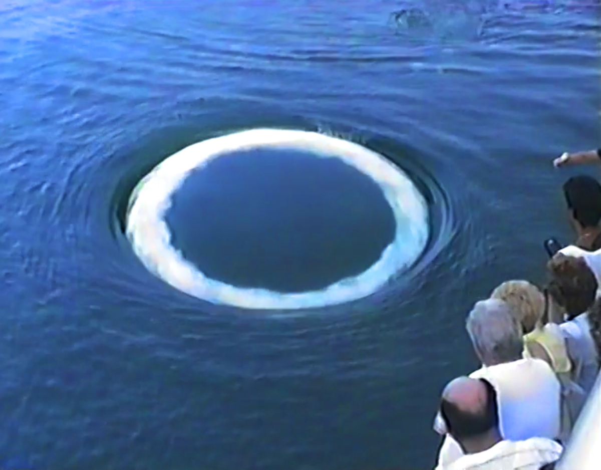 video still of people on a boat looking at w white ring on the ocean